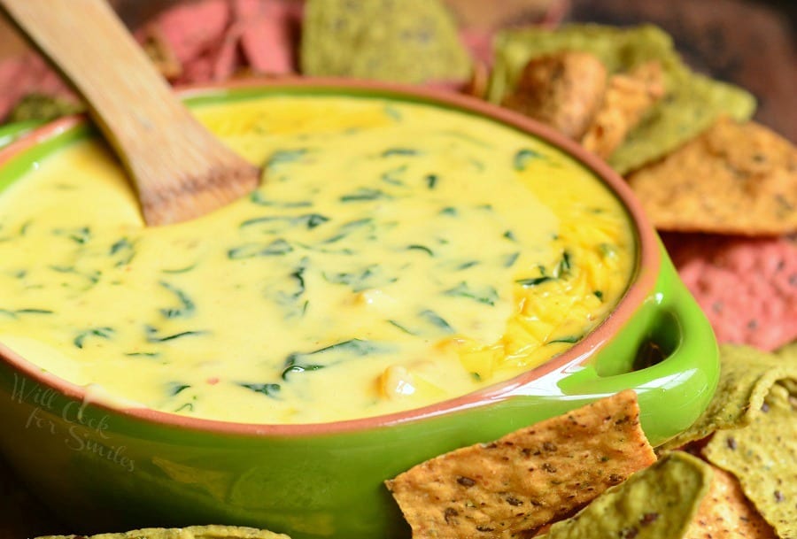 Shrimp and Spinach Queso Dip in a green bowl with a wooden spoon in it and tortilla chips around it 