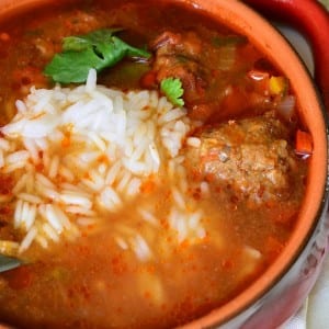 Red clay crock filled with mexican meatballs soup on a white cloth with a spoon behind the crock to the right with a spoon in the soup as viewed from above
