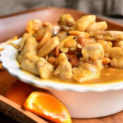 white decorative bowl filled with nutty light orange chicken on a wooden serving tray with sliced oranges around the bowl