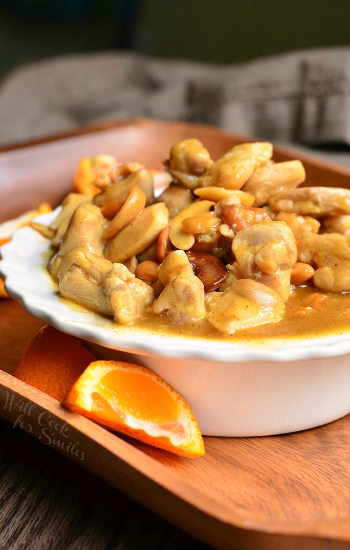 white decorative bowl filled with nutty light orange chicken on a wooden serving tray with sliced oranges around the bowl