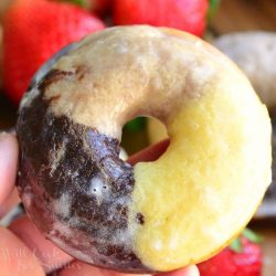 Baked napoleon glazed doughnuts glazed on a wooden cuttingboard with strawberries in the background and a hand hold one doughnut above the rest