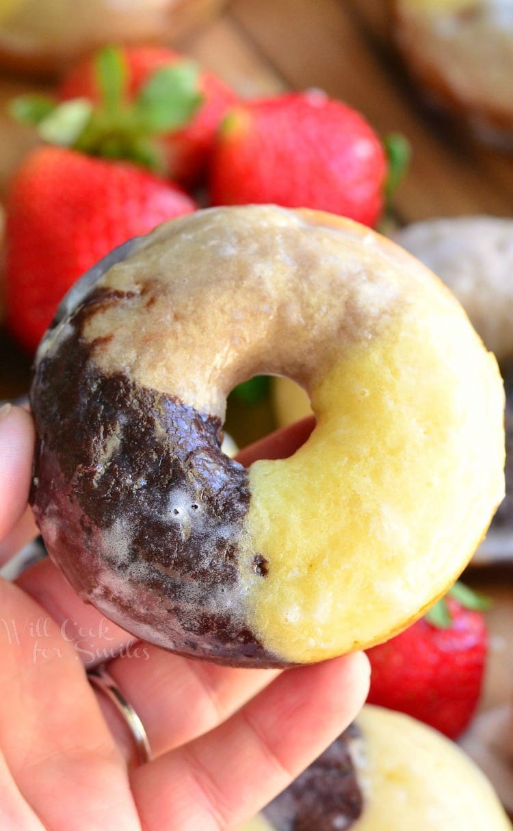 Baked napoleon glazed doughnuts glazed on a wooden cuttingboard with strawberries in the background and a hand hold one doughnut above the rest