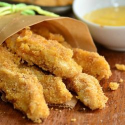 Wax paper holder holding easy honey mustard baked chicken fries on a wooden table with a small white saucer in the background filled with sauce and a pan with additional chicken fries in the background to the left as viewed close up