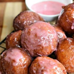 pile of strawberry glazed doughnuts on a wire cooling rack with a small white dish and dipping sauce in the background