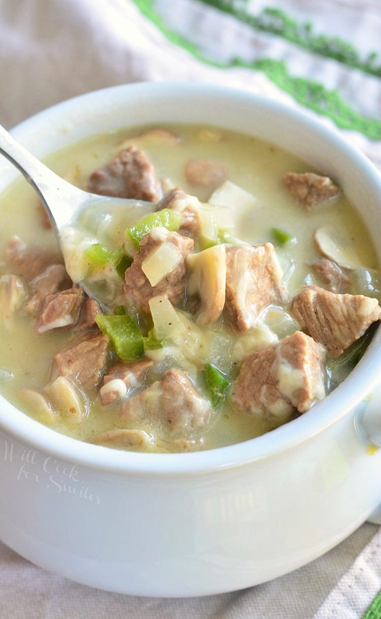 white crock filled with steak and cheese soup on a wooden table with a spoon holding up one bite of soup above the crock