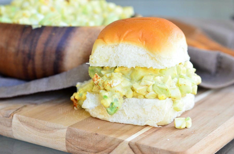 Avocado Cucumber Egg Salad on a bun sitting on a cutting board with a wood bowl of egg salad in the background on 