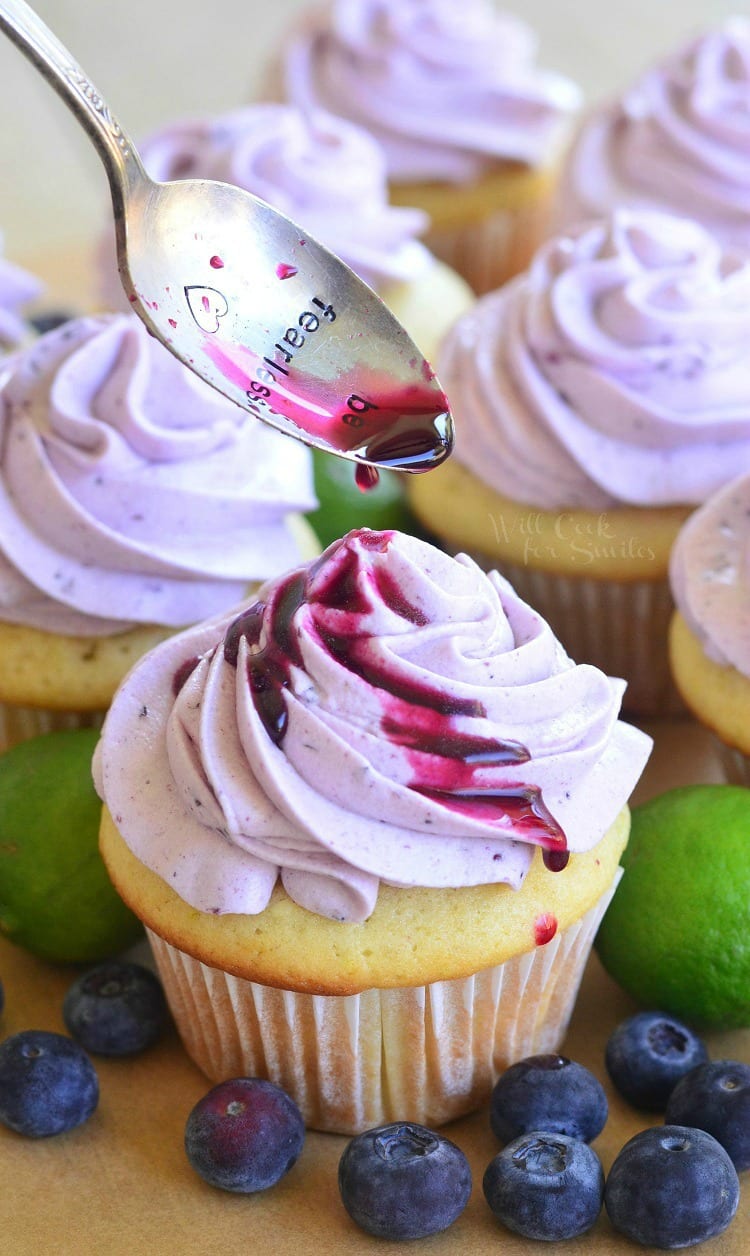 spooning sauce over cupcakes with Fresh Blueberry Cream Cheese Frosting on a cutting board with blueberries and limes 