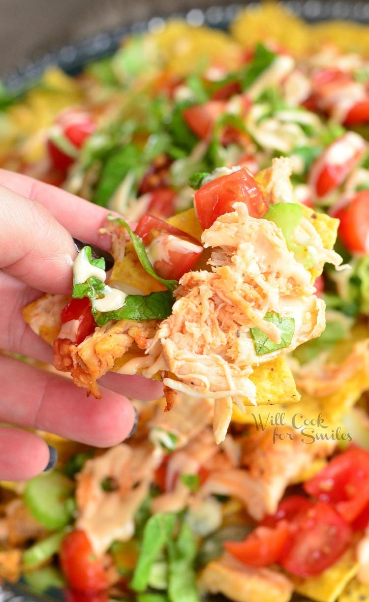 holding a nacho and Buffalo Chicken Nachos on a metal serving plater on a table in the background 