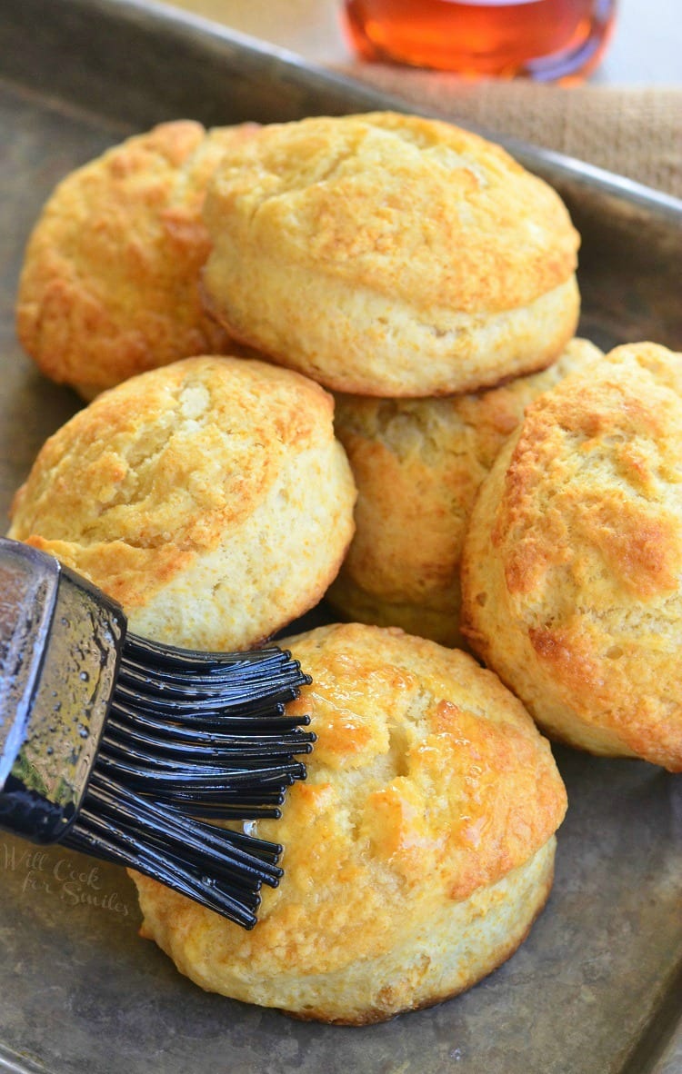 Honey Butter Biscuits stacked on a baking sheet brushing butter on one of them 