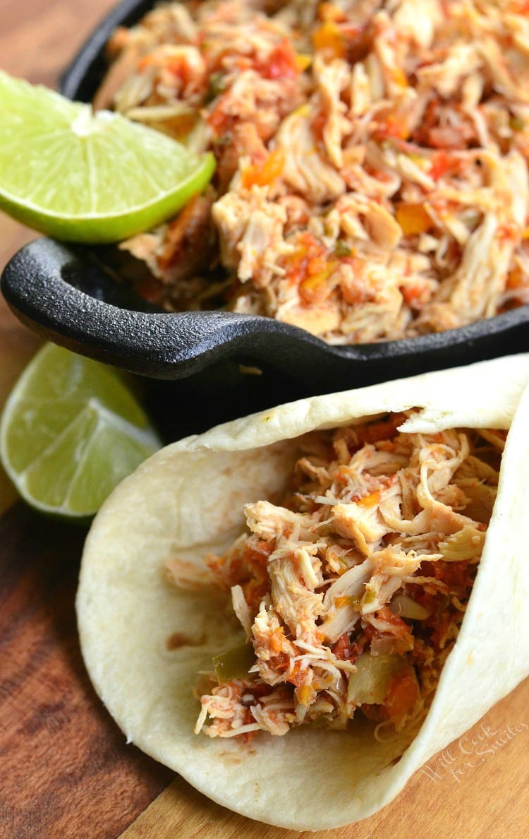 Close up view of crock pot shredded salsa chicken on a black skillet with a lime garnish on a wooden cutting board with some chicken wrapped in a small tortilla