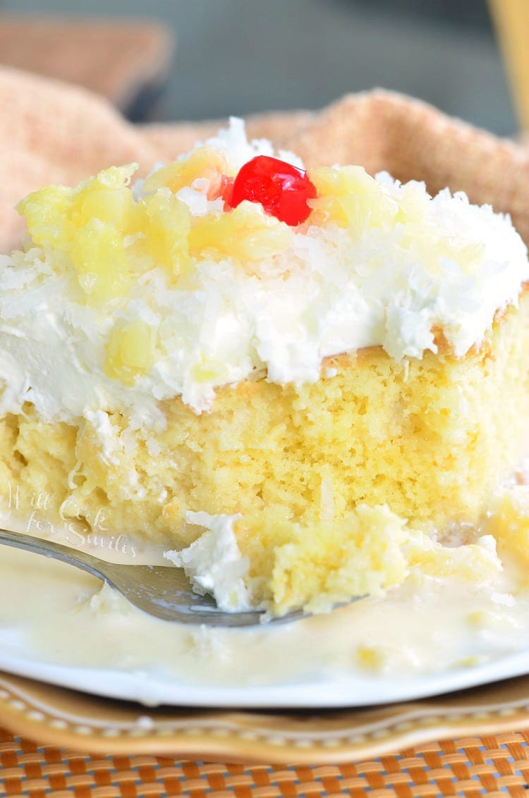 a slice of Tres Leches Cake with white coconut frosting, pineapple and a cherry on top with a fork with some cake on it 