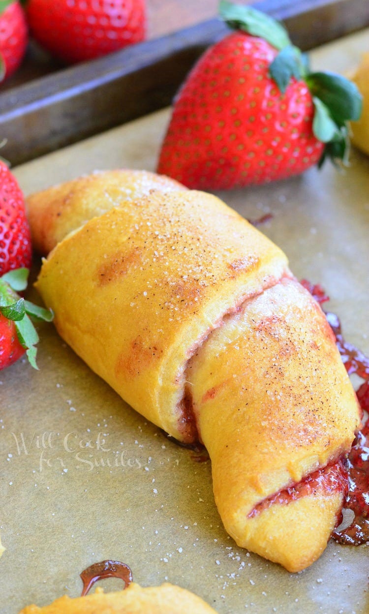crescent rolls with strawberry jam inside them on a baking shit with parchment paper and strawberries in the background