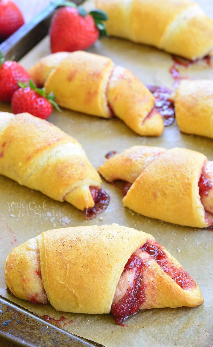 6 strawberry crecent rolls on a baking sheet tray with strawberries scattered around the tray