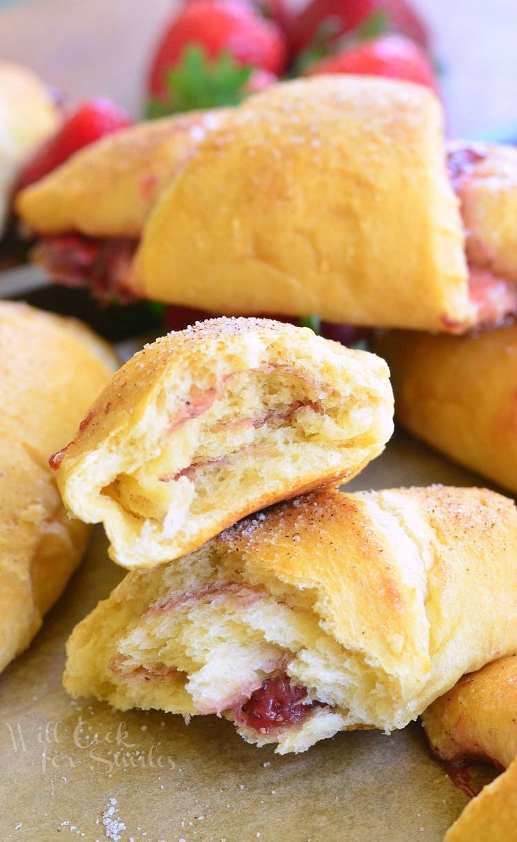 crescent rolls with strawberry jam inside them on a baking shit with parchment paper and strawberries in the background with a crescent roll torn in half