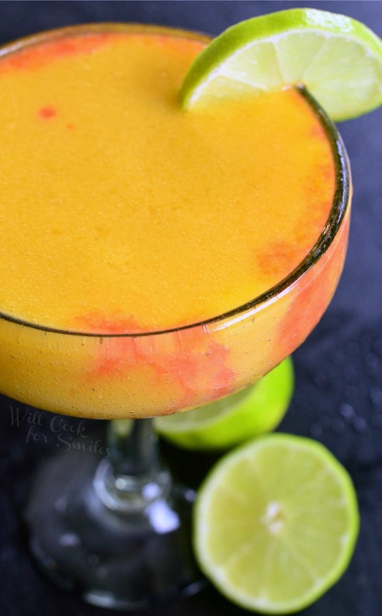 view from above of a sunset margarita garnished with lime while sliced limes rest on a black table by the glass' stem