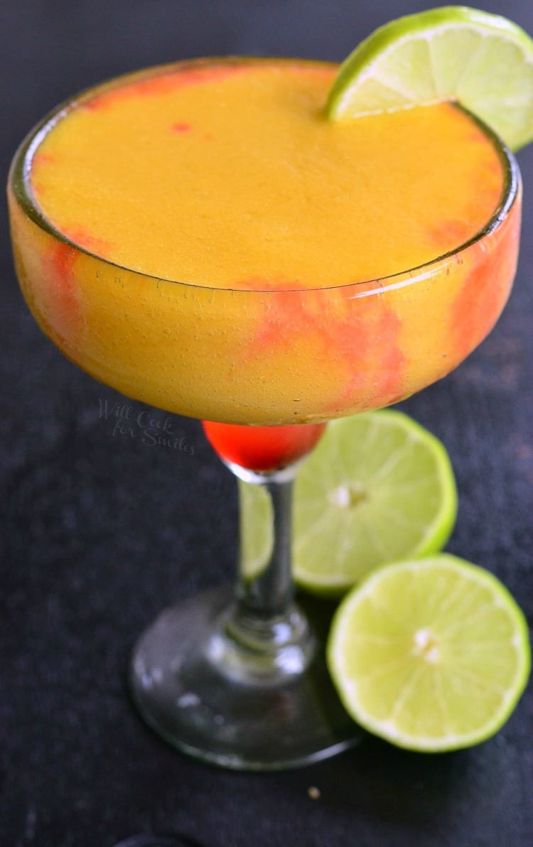 a sunset margarita garnished with lime while sliced limes rest on a black table by the glass' stem