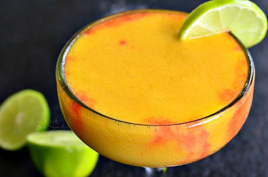 view from above and close up of a sunset margarita garnished with lime while sliced limes rest on a black table by the glass' stem