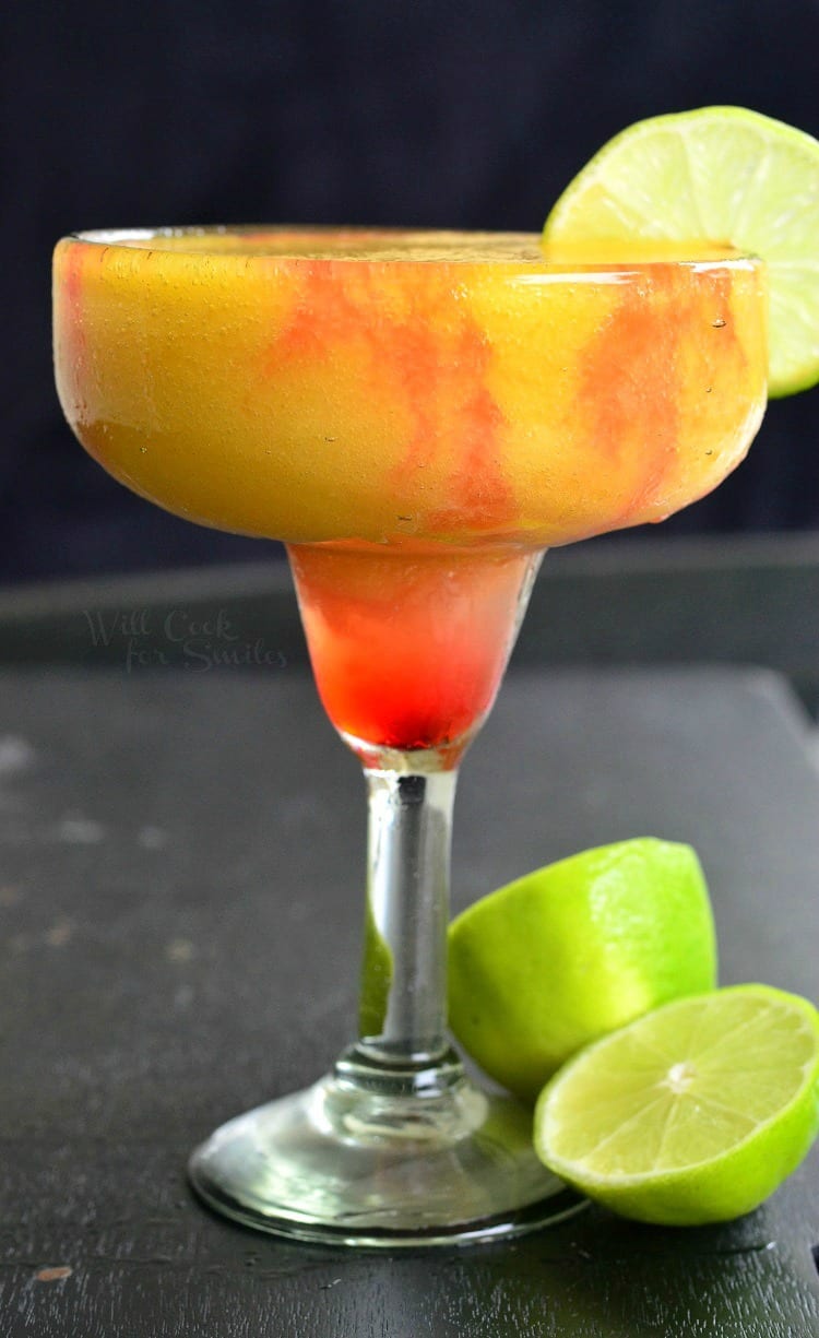 A sunset margarita garnished with lime while sliced limes rest on a black table by the glass' stem