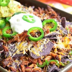 Baking sheet pan with pulled pork nachos topped with sour cream and sliced jalapenos on a wooden table with a white and yellow cloth in the background