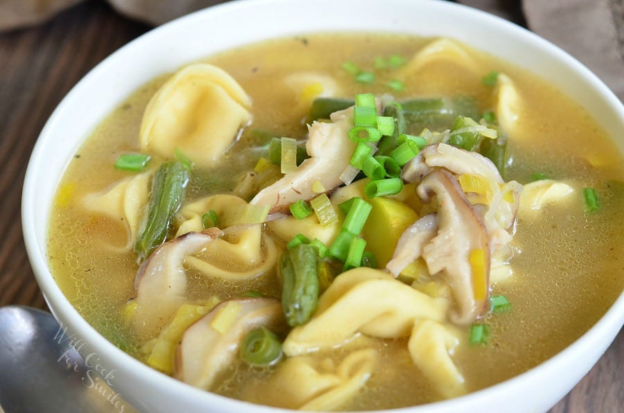 horizontal image of Vegetable Medley Tortellini Soup in a white bowl with green onion as garnish and a spoon to the left of the bowl 