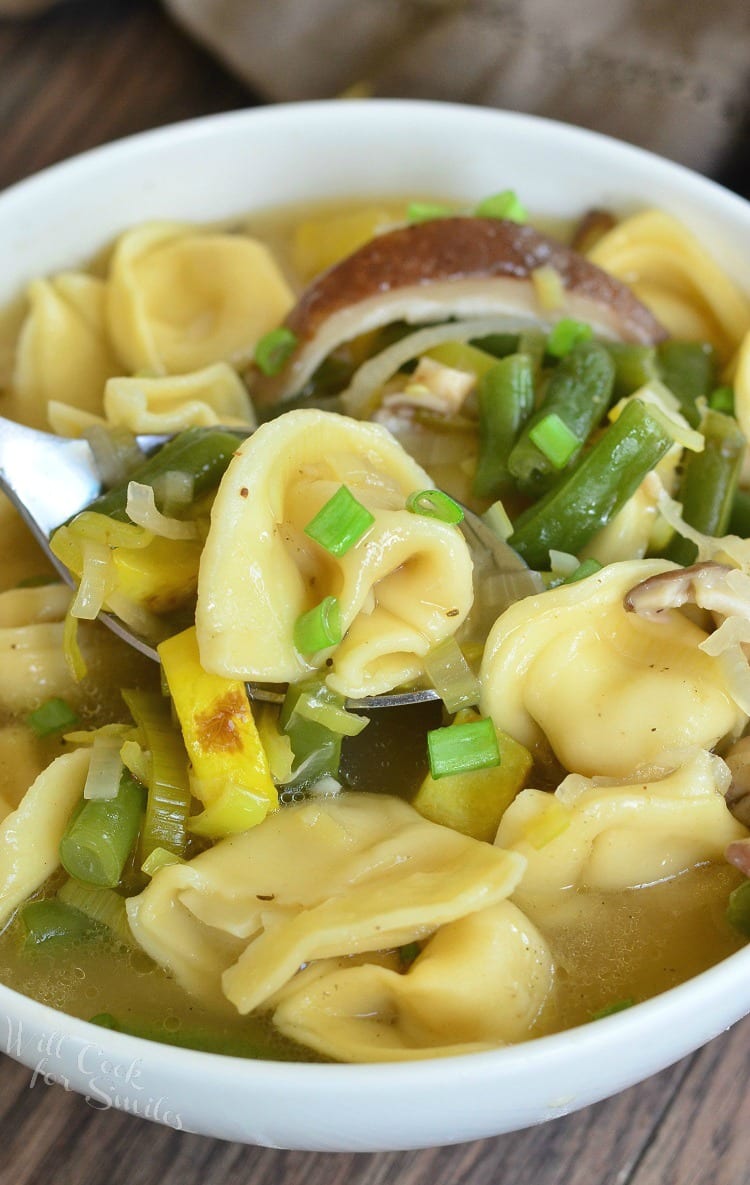 Vegetable Medley Tortellini Soup in a white bowl with green onion as garnish and a spoon lifting some out of the bowl 