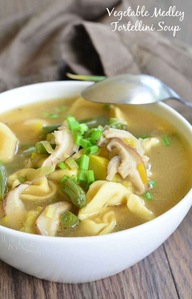 Vegetable Medley Tortellini Soup in a white bowl with green onion as garnish and a spoon over top of the bowl 