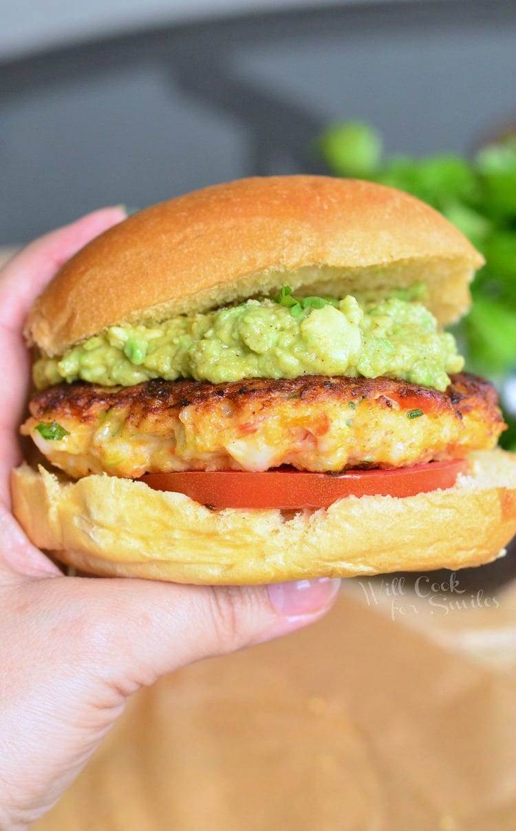 holding Avocado Shrimp Burgers with guacamole on top and tomato on a bun 