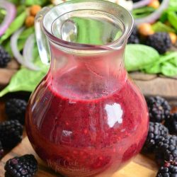 Glass of black berry poppy vinaigrette on a wooden board with blackberries scattered around the base of glass and a salad in the background