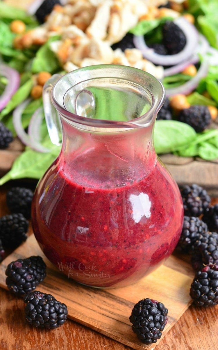 Blackberry Poppy Seed Vinaigrette in a glass container with blackberries around it and salad in the background on a cutting board 