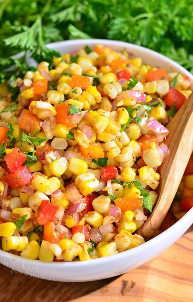 Cajun Corn Salad with corn, red and orange bell peppers, red onion, an cilantro in a white bowl with a wooden spoon 