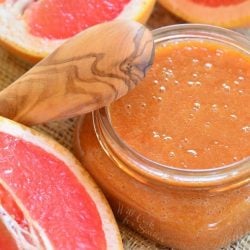Glass jar filled with homemade grapefruit syrup and sauce on a tan placemat with a wooden spoon leaning on the jar and sliced grapefruit laying around the jar as viewed from above.