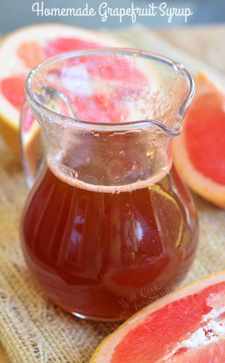 Glass jar filled with homemade grapefruit syrup and sauce on a tan placemat with a wooden spoon leaning on the jar and sliced grapefruit laying around the jar.