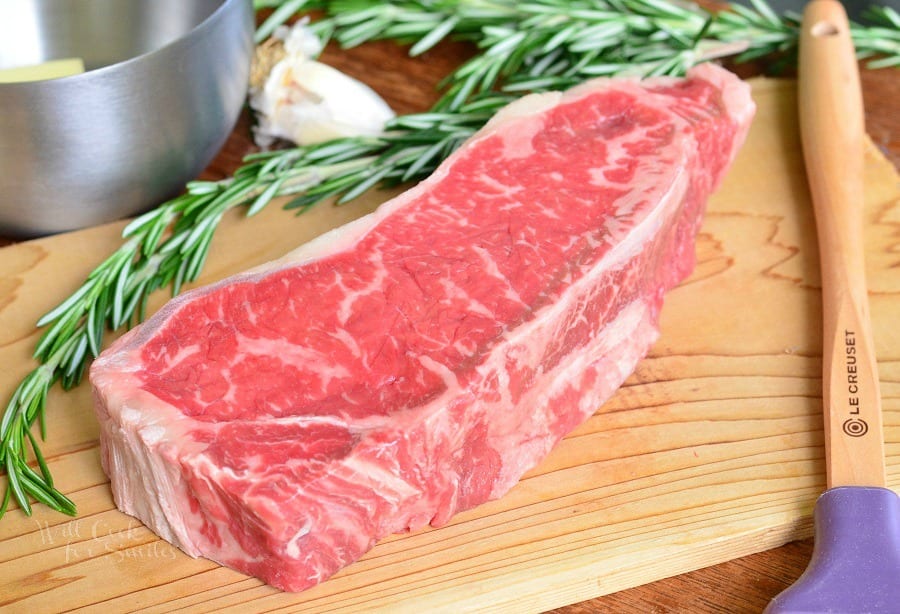 raw steak on the cutting board with rosemary and spatula next to it