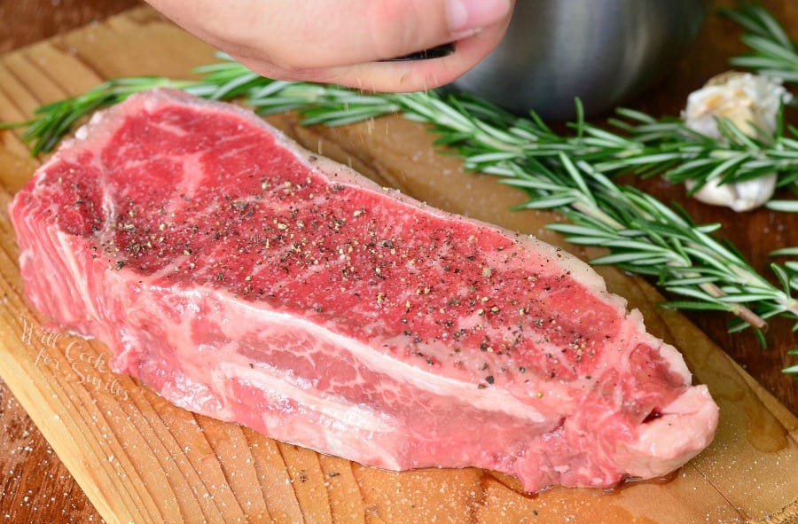 grinding black pepper over raw steak on the cutting board with rosemary sprig next to it.