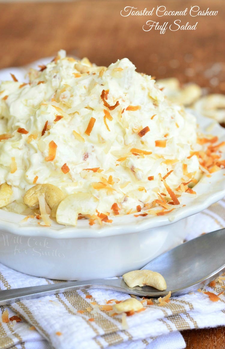 Toasted Coconut and Cashews on top of Fluff Salad in a white bowl on a white and brown kitchen towel with a spoon across the front 