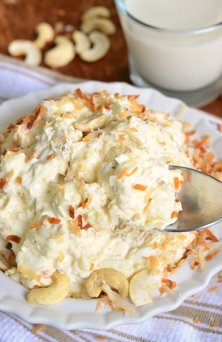 top view of Toasted Coconut and Cashews on top of Fluff Salad in a white bowl on a white and brown kitchen towel with a spoon getting some out