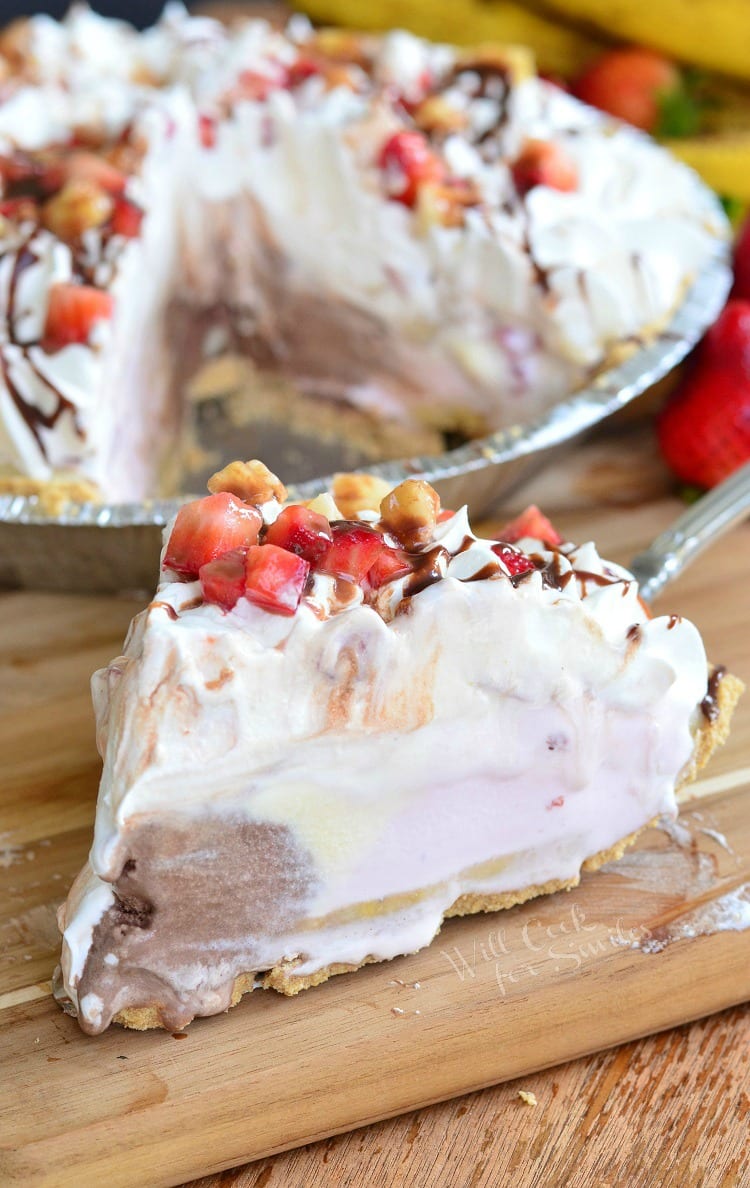 A slice of Banana Split Ice Cream Pie with whipped cream, strawberries, banana, and chocolate syrup on top on a table 