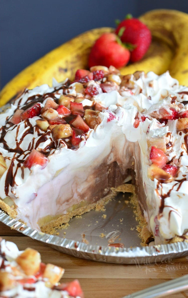 Banana Split Ice Cream Pie in a disposable metal pie pan with whipped cream, strawberries, banana, and chocolate syrup on top with a slice taken out 