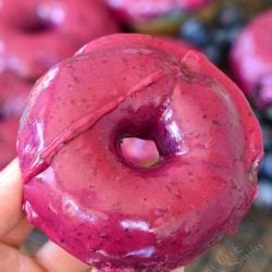 A hand holding a Blueberry doughnuts with blueberry cream whiskey glaze on a wire rack with blueberries scattered around them