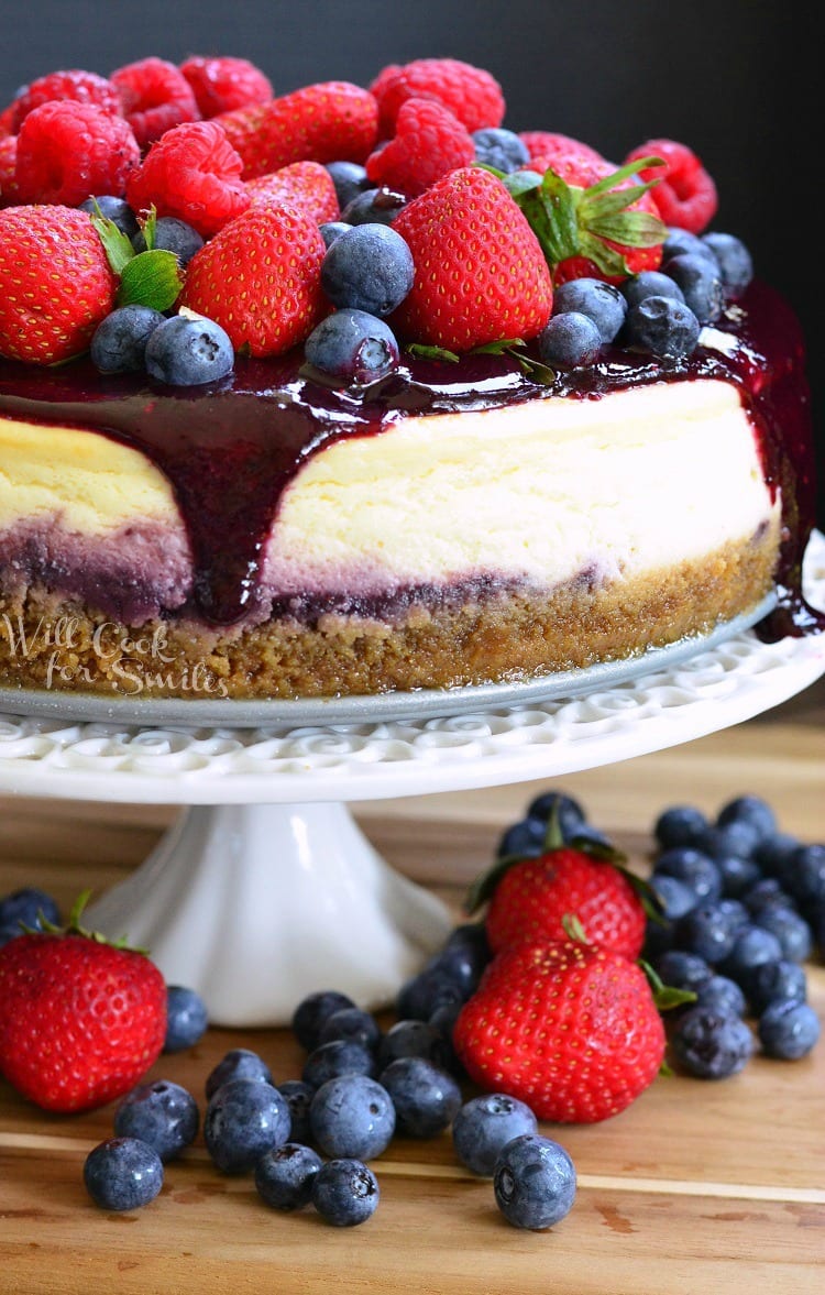 cheesecake with berry sauce, strawberries, blueberries, and raspberries on top on a white cake stand with blue berries and strawberries under it on a wood table