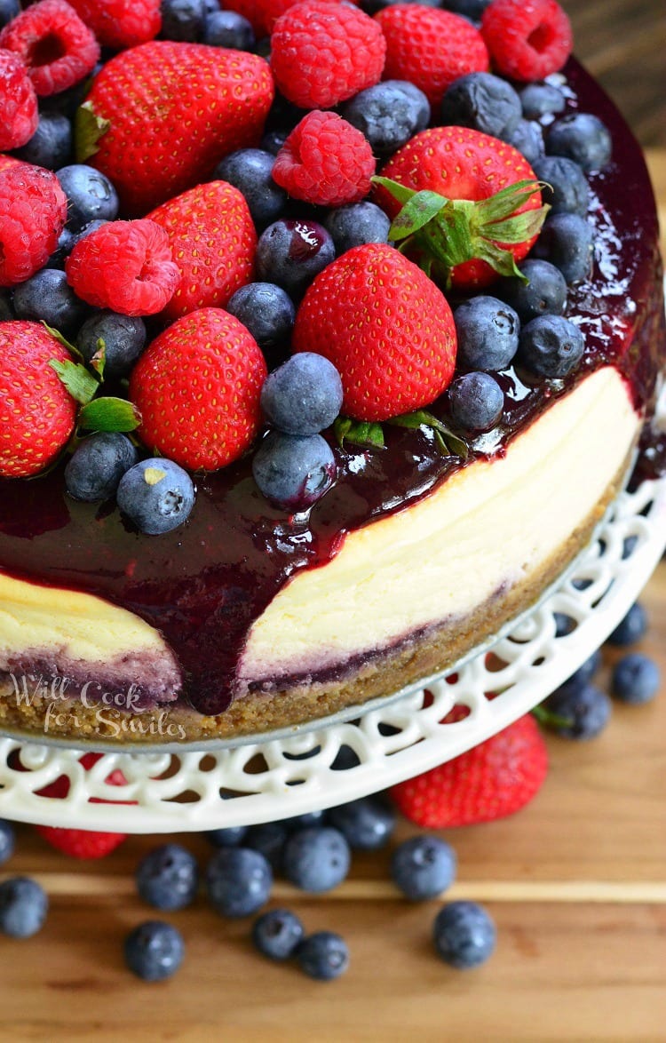 top view photo of cheesecake with berry sauce, strawberries, blueberries, and raspberries on top on a white cake stand with blue berries and strawberries under it on a wood table