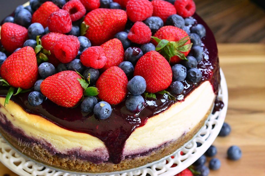 cheesecake with berry sauce, strawberries, blueberries, and raspberries on top on a white cake stand with blue berries and strawberries under it on a wood table