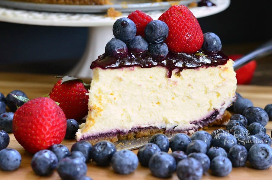 slice of cheesecake with berry sauce, raspberries, and strawberries on top on a cutting board with strawberries and blueberries around it