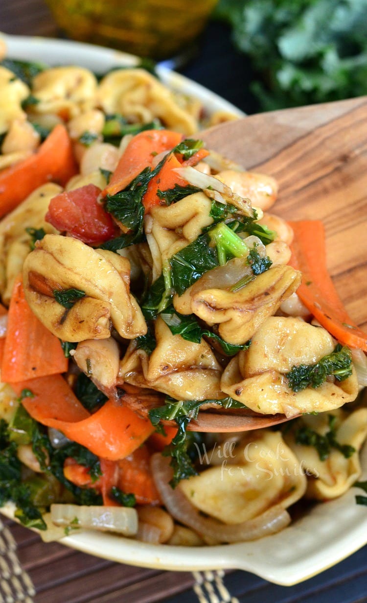 Tortellini pasta with carrots, onions, tomato in a yellow bowl with a wooden spoon scooping some out 