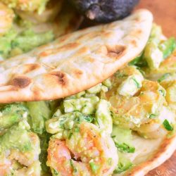 2 avocado shrimp flatbread sandwiches laying on a wooden cutting board with 2 avocados in the background