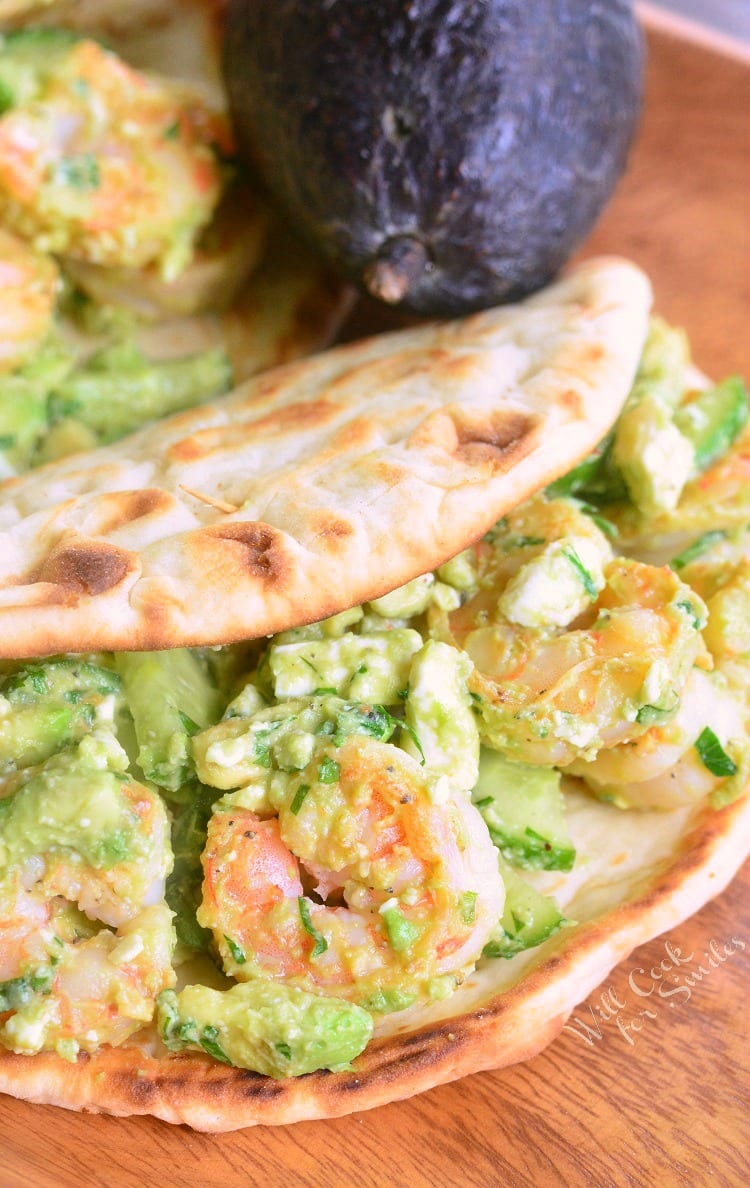 2 avocado shrimp flatbread sandwiches laying on a wooden cutting board with 2 avocados in the background