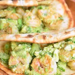 2 avocado shrimp flatbread sandwiches laying on a wooden cutting board with 2 avocados in the background