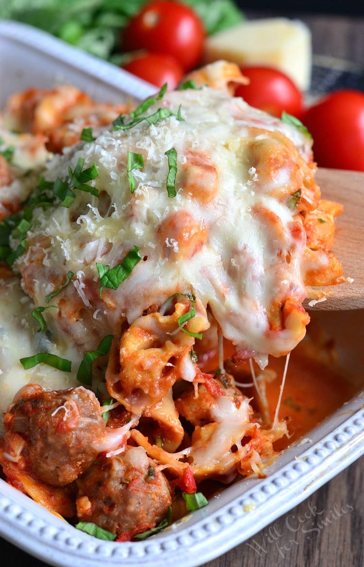 Chicken, Sausage, and pasta with cheese on top in a casserole dish with a wooden spoon lifting some out 