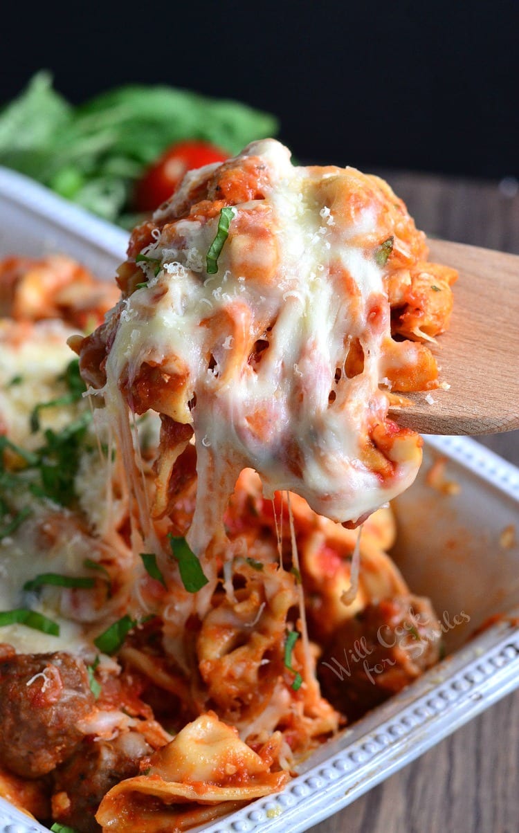 Chicken, Sausage, and pasta with cheese on top in a casserole dish with a wooden spoon lifting some out 