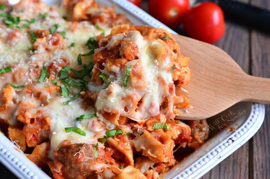 Chicken, Sausage, and pasta with cheese on top in a casserole dish with a wooden spoon lifting some out 